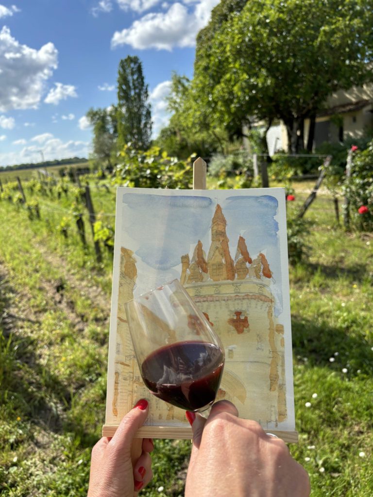 Une main tient un verre de vin rouge devant une aquarelle peinte au vin. Cette activité proposée par Laetitia Ouspointour s'inspire d'artistes comme Christina LoCascio et Elisabetta Rogai qui ont exploré le vin comme une matière pour peindre.